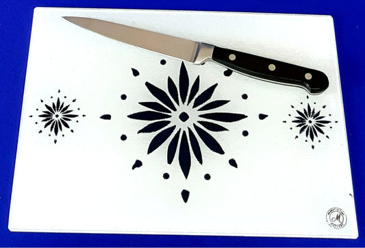White cutting board with blue flowerburst design. A chopping knife on top of the board. Board displayed on a blue background. Premium glass worktop saver and serving board. A4 size.