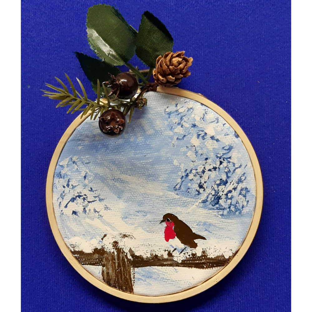 Original signed artwork, Circle Christmas robin canvas decoration. Robin sitting on a snow covered fence with frosty shades of blue background. Sealed with a varnish finish, dusted with a hint of sparkle and added festive trimmings. Framed in a sustainable bamboo hoop ring.
