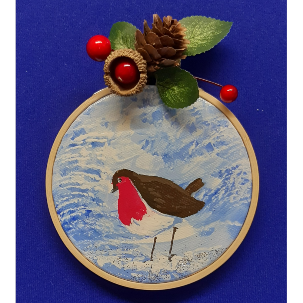 Original signed artwork, Circle Christmas robin canvas decoration. Robin in the centre looking to the left on the snow covered ground, with frosty shades of blue background. Sealed with a varnish finish, dusted with a hint of sparkle and added festive trimmings. Framed in a sustainable bamboo hoop ring.