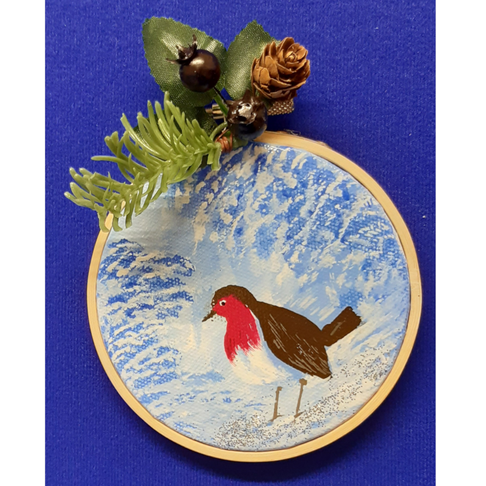 Original signed artwork, Circle Christmas robin canvas decoration. Robin on snow covered ground looking to the left, with frosty shades of blue background. Sealed with a varnish finish, dusted with a hint of sparkle and added festive trimmings. Framed in a sustainable bamboo hoop ring.