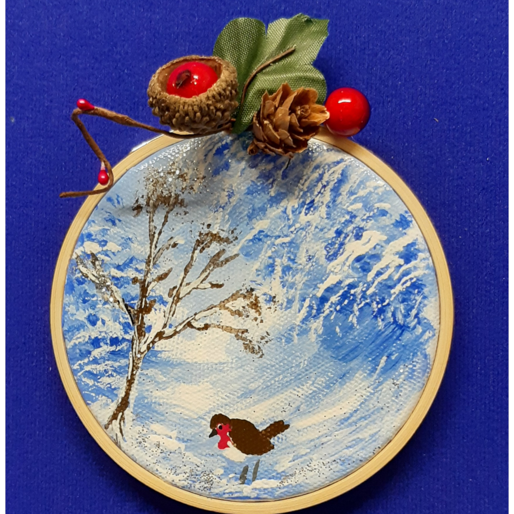 Original signed artwork, Circle Christmas robin canvas decoration. Robin on snow covered ground looking to the left, with frosty shades of blue tree background. Sealed with a varnish finish, dusted with a hint of sparkle and added festive trimmings. Framed in a sustainable bamboo hoop ring.
