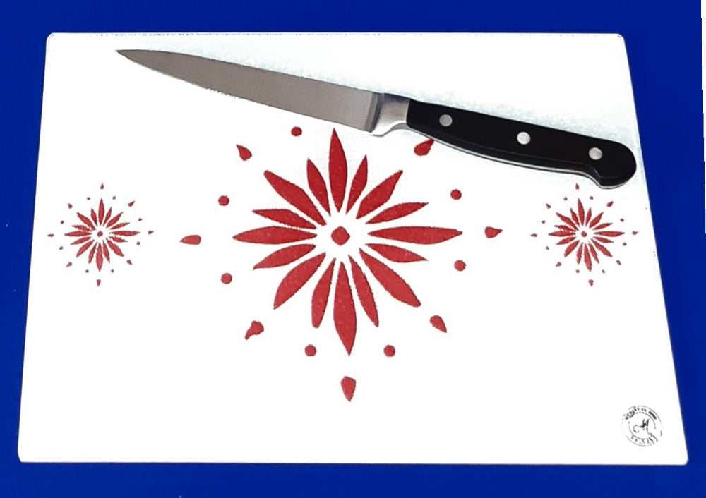 White cutting board with red flowerburst design. A chopping knife on top of the board. Board displayed on a blue background. Premium glass worktop saver and serving board. A4 size.
