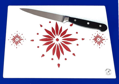 White cutting board with red flowerburst design. A chopping knife on top of the board. Board displayed on a blue background. Premium glass worktop saver and serving board. A4 size.