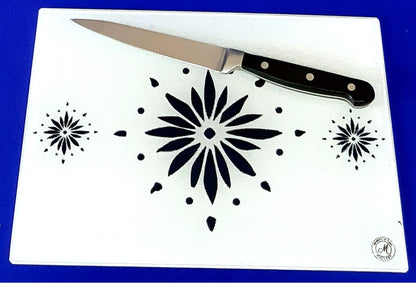 White cutting board with blue flowerburst design. A chopping knife on top of the board. Board displayed on a blue background. Premium glass worktop saver and serving board. A4 size.