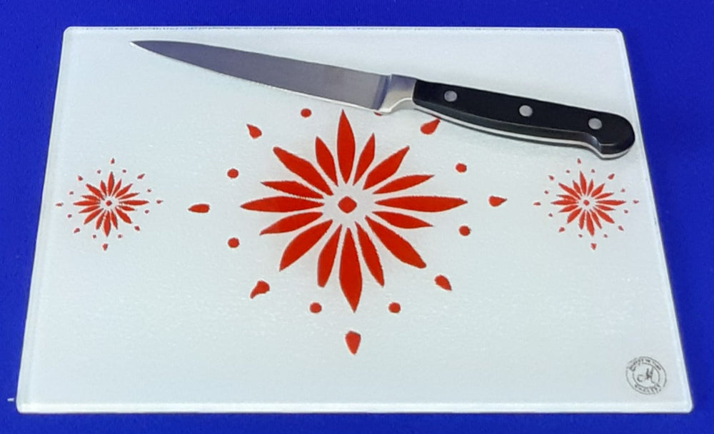 White cutting board with red flowerburst design. A chopping knife on top of the board. Board displayed on a blue background. Premium glass worktop saver and serving board. A4 size.